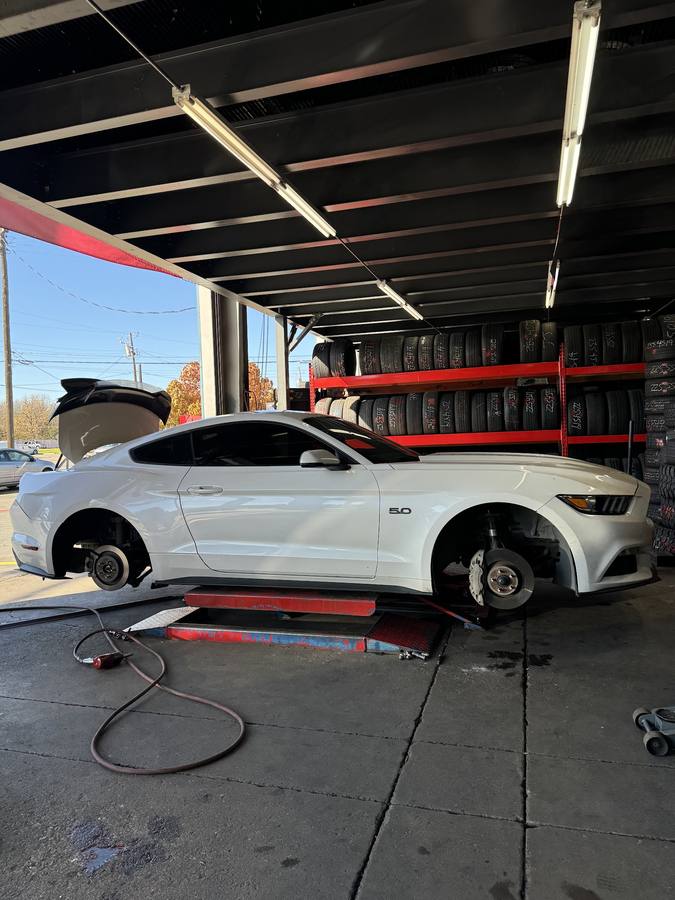 A white Mustang raised on a lift with its wheels removed, tire racks behind it.
