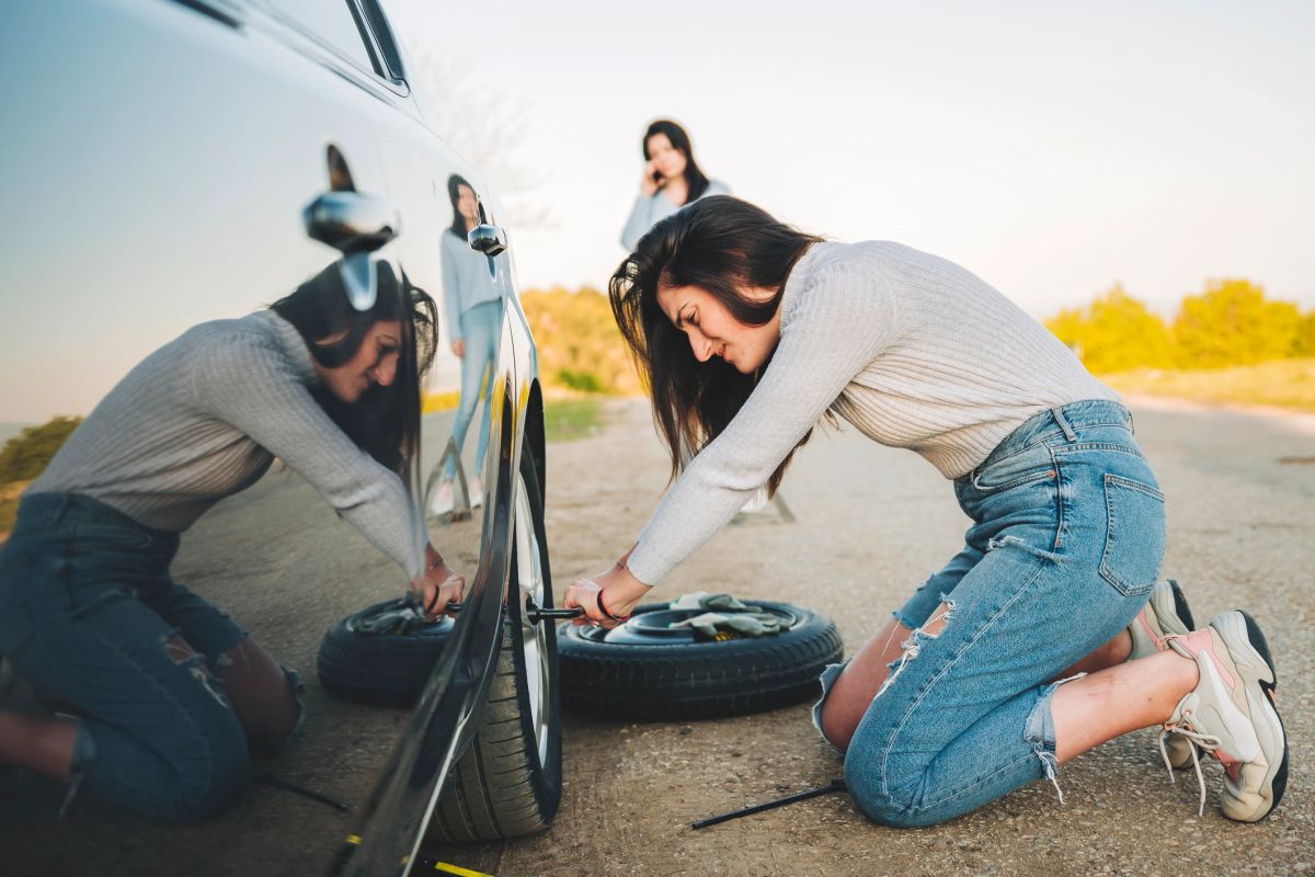 A woman kneeling beside a car changing a flat tire on the roadside.